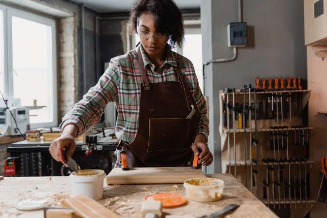 Une femme menuisier portant un tablier en cuir applique de la colle à bois sur une pièce de bois dans son atelier.